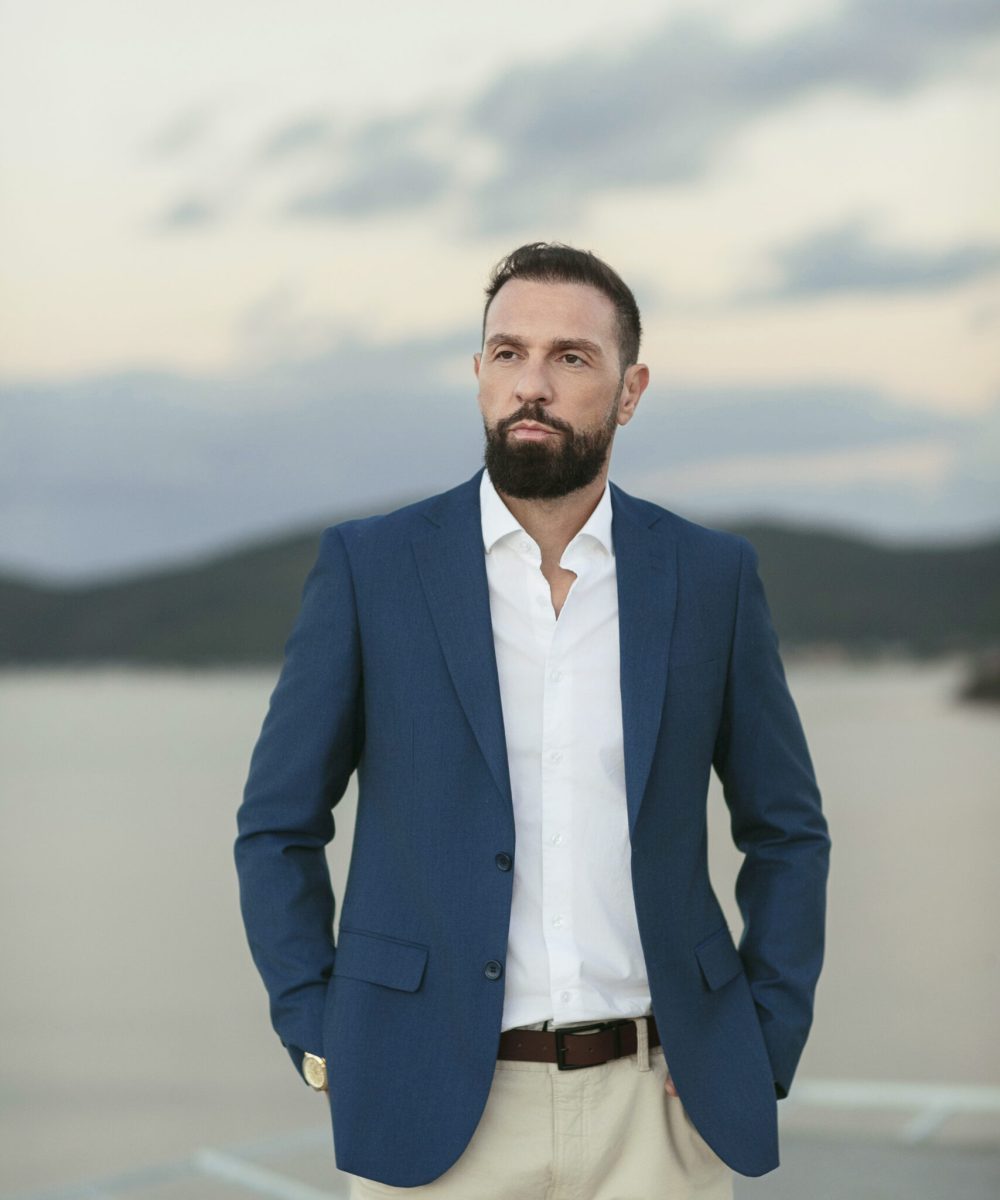 Bruno Mesko Dias, advogado e cofundador da Ágata Imóveis, posando à beira-mar em Porto Belo com traje social, transmitindo elegância e profissionalismo.